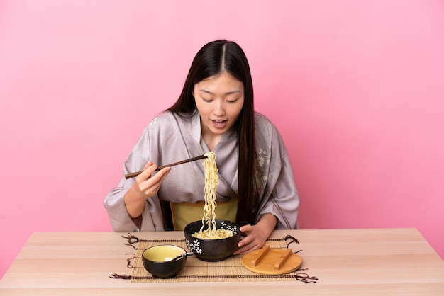 Junge chinesische Frau, die Kimono trägt und Nudeln in einem Tisch über rosa Wand isst