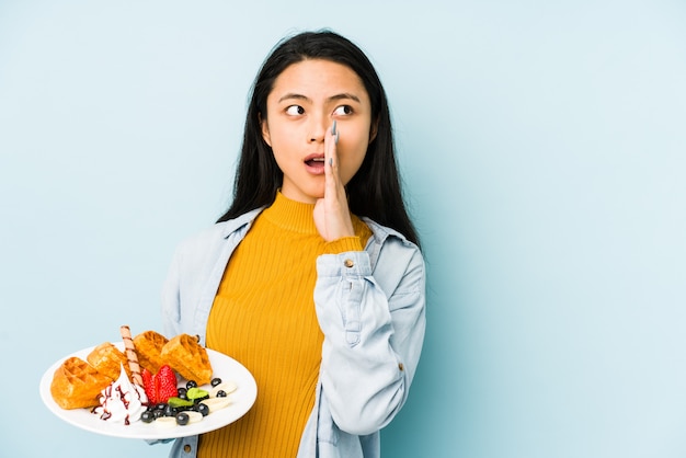 Junge chinesische Frau, die Waffel auf blauer Wand hält, die Tempel mit Finger zeigt, denkend, auf eine Aufgabe fokussiert.