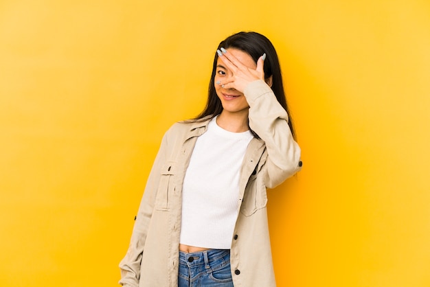 Junge chinesische Frau isoliert auf einer gelben Wand blinzeln durch Finger, verlegenes bedeckendes Gesicht.