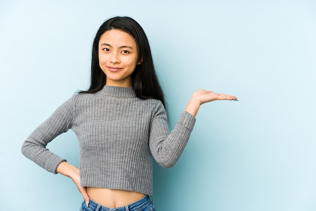 Junge chinesische Frau lokalisiert auf einer blauen Wand, die einen Kopienraum auf einer Handfläche zeigt und eine andere Hand auf Taille hält.