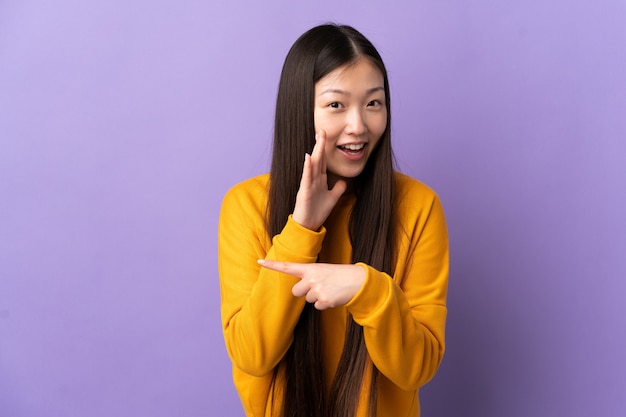 Junge chinesische Frau über isolierter lila Wand, die zur Seite zeigt, um ein Produkt zu präsentieren und etwas zu flüstern