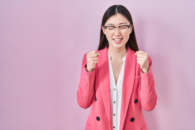 Junge chinesische Geschäftsfrau mit Brille freut sich über den Erfolg mit erhobenen Armen und geschlossenen Augen und feiert den Sieg lächelnd. Gewinner-Konzept.