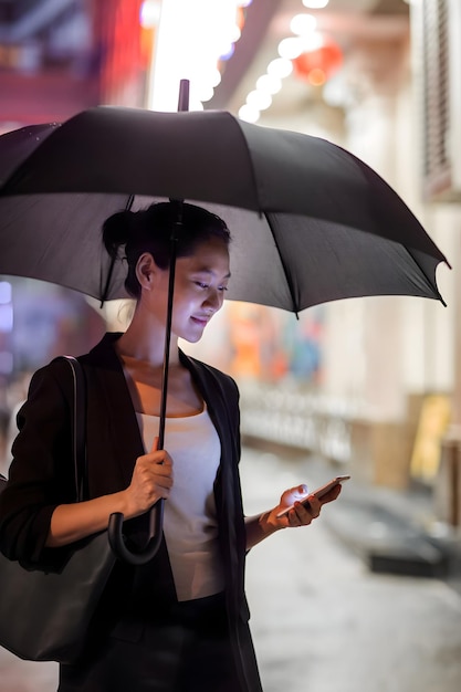 Junge chinesische Geschäftsfrau mit einem Regenschirm wartet auf der Straße und benutzt ein Telefon in Hongkongs Yau Ma Tei