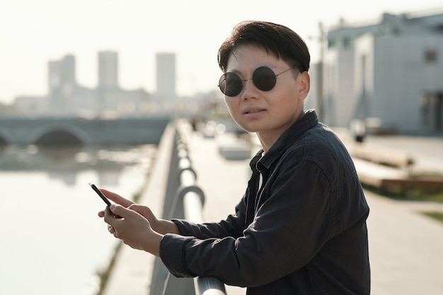 Junge chinesische Geschäftsfrau mit Sonnenbrille, die am Flussufer steht und im Smartphone scrollt