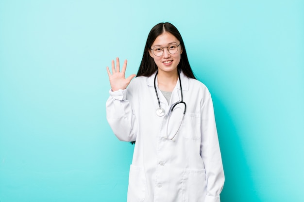 Junge chinesische Ärztin, die fröhlich lächelt und Nummer fünf mit den Fingern zeigt.