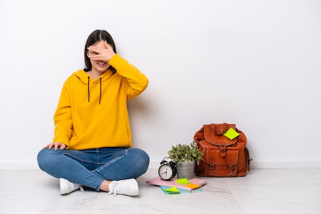 Junge chinesische Studentin, die auf dem Boden sitzt, isoliert auf einer weißen Wand, die die Augen mit den Händen bedeckt und lächelt