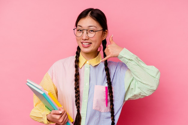 Junge chinesische Studentin, die Bücher hält, die ein Mode-Mehrfarbenhemd und -geflecht tragen, lokalisiert auf rosa Hintergrund, der eine Handy-Anrufgeste mit den Fingern zeigt.