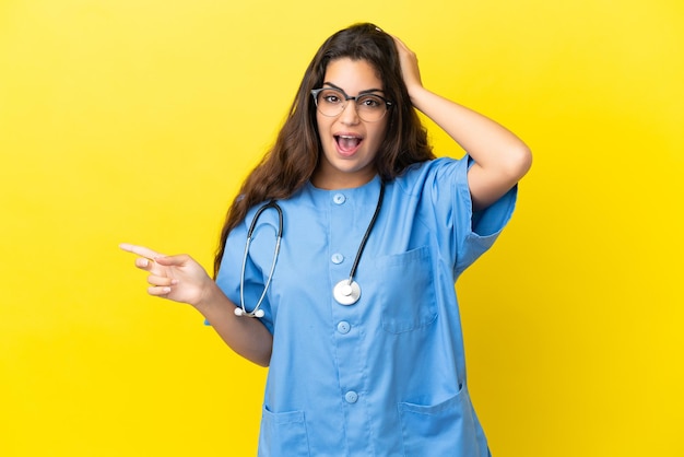 Junge Chirurg Arzt Frau isoliert auf gelbem Hintergrund überrascht und zeigt mit dem Finger zur Seite