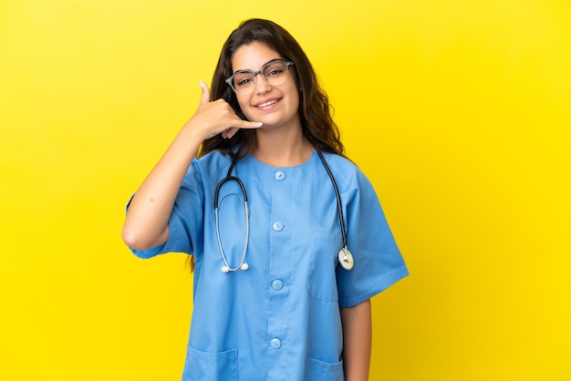 Junge Chirurgdoktorfrau lokalisiert auf gelbem Hintergrund, der Telefongeste macht. Ruf mich zurück Zeichen