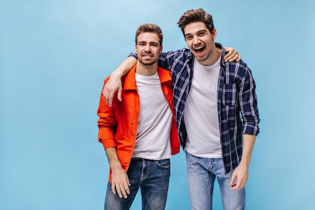 Junge coole bärtige Männer in orangefarbener Jacke, kariertem Hemd und Jeanslächeln schauen in die Kamera und umarmen sich auf blauem Hintergrund