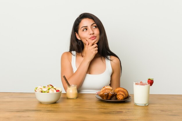 Junge curvy Frau, die ein Frühstück seitlich schaut mit zweifelhaftem und skeptischem Ausdruck nimmt.