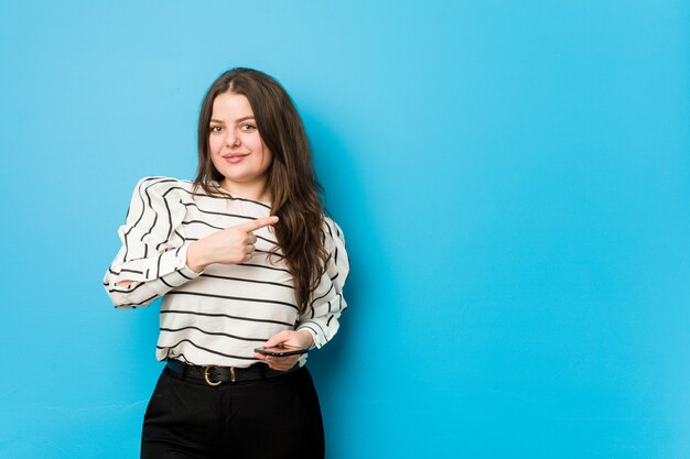 Junge curvy Frau, die ein Telefon lächelt und beiseite zeigt und etwas auf Leerstelle hält.