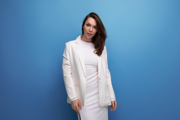 Junge Dame des Modegeschäfts im weißen Kleid und in der Jacke, die auf Studiohintergrund mit Exemplar aufwirft