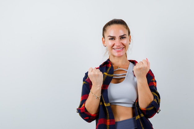 Junge Dame, die Gewinnergeste in Top, kariertem Hemd zeigt und glücklich aussieht. Vorderansicht.