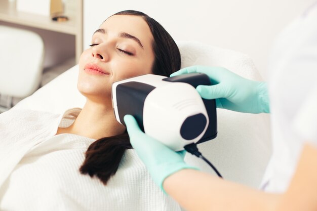 Junge Dame, die ihre Augen schließt und lächelt, während eine erfahrene Kosmetikerin eine Hautanalysekamera in der Nähe ihres Gesichts hält