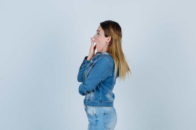 Junge Dame, die im Denim-Outfit die Hand auf dem Mund hält und schockiert aussieht. .