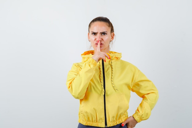 Junge Dame, die Schweigegeste in gelber Jacke zeigt und ernst aussieht. Vorderansicht.