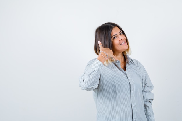 Junge Dame, die Telefongeste in übergroßem Hemd zeigt und selbstbewusst aussieht. Vorderansicht.