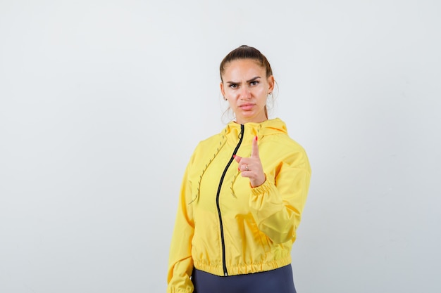 Junge Dame droht mit dem Finger in gelber Jacke und sieht ernst aus. Vorderansicht.