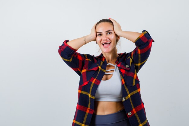 Junge Dame in Top, kariertes Hemd mit den Händen auf dem Kopf und fröhlich aussehend, Vorderansicht.