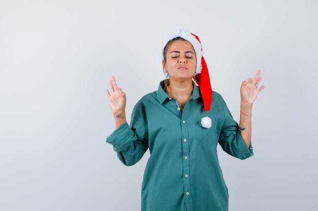 Junge Dame in Weihnachtsmütze, Hemd, das Meditationsgeste zeigt und entspannt aussieht, Vorderansicht.