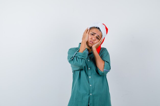 Junge Dame mit den Händen auf den Wangen in Weihnachtsmütze, Hemd und enttäuscht, Vorderansicht.