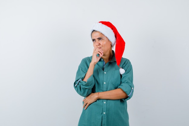 Junge Dame mit der Hand auf dem Mund in Weihnachtsmütze, Hemd und nachdenklich aussehend. Vorderansicht.