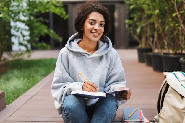 Foto junge dame mit dunklem lockigem haar sitzt und macht notizen im notizbuch, während glücklich