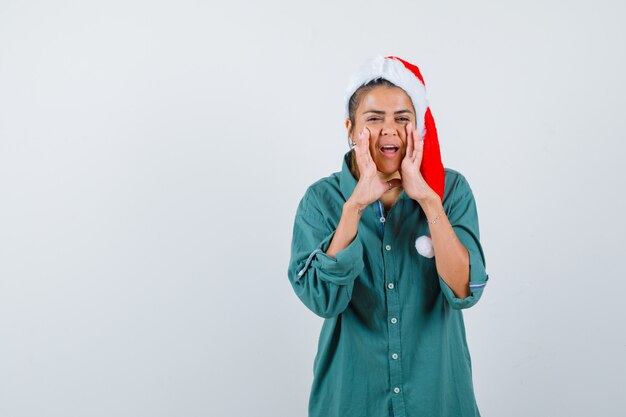 Junge Dame mit Händen in der Nähe des Mundes, um Geheimnis in Weihnachtsmütze, Hemd und fröhlichem Aussehen zu erzählen, Vorderansicht.