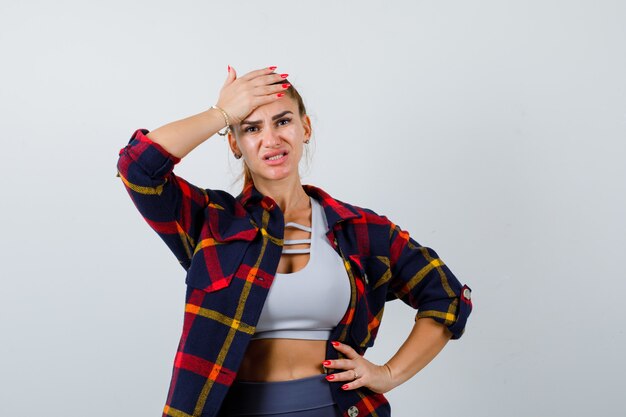 Junge Dame mit Hand auf der Stirn im Oberteil, kariertem Hemd und vergesslich aussehend. Vorderansicht.