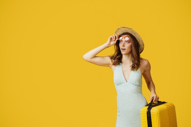 Junge Dame mit Sonnenbrille und Sommerhut, die Koffer hält und Kopienraum auf gelbem Studiohintergrund betrachtet