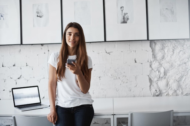 Junge Dame sitzt im Coworking Center mit Smartphone und Laptop und schaut lächelnd in die Kamera