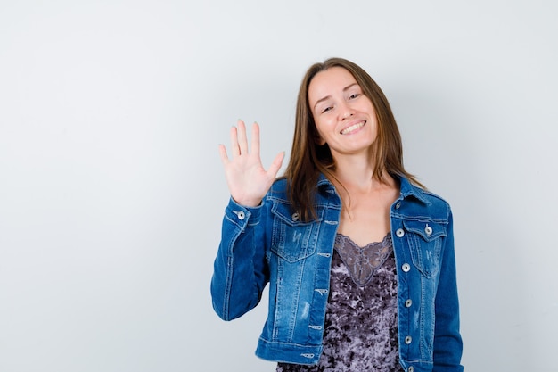 Junge Dame winkt mit der Hand zur Begrüßung in Bluse, Jeansjacke und sieht fröhlich aus, Vorderansicht.