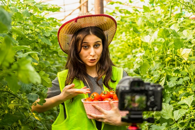 Junge Dame zeigte mit der Hand auf die Tomaten und sprach mit der Kamera im Gewächshaus