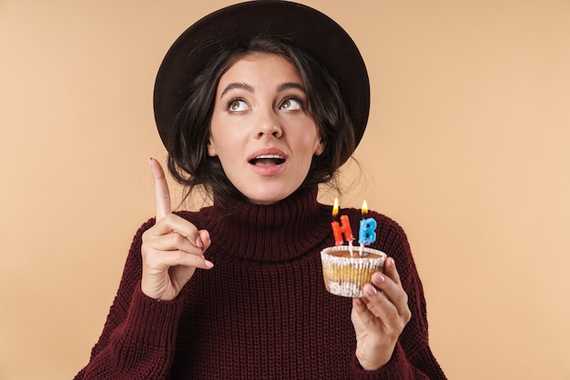junge denkende brünette Frau isoliert über beige Wand Wand hält Cupcake mit alles Gute zum Geburtstag Kerzen zeigen.