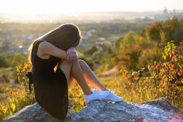 Junge depressive Frau im schwarzen kurzen Sommerkleid, das auf einem Felsen sitzt, der draußen bei Sonnenuntergang denkt. Modische Frau, die im warmen Abend in der Natur betrachtet.