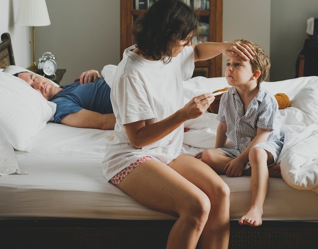 Junge, der auf dem Bett und seiner Mutter überprüft Temperatur sitzt