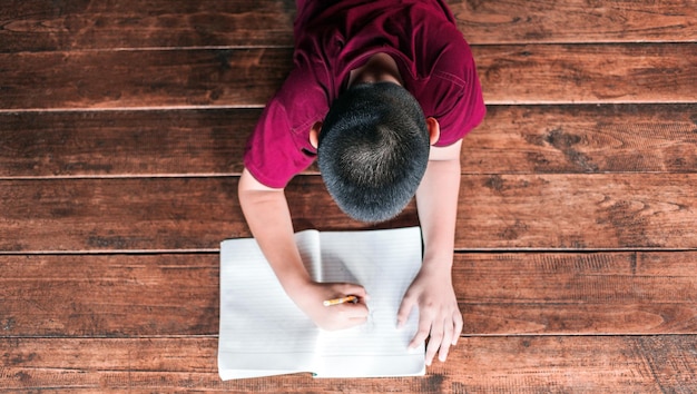 Junge, der auf dem Holzboden sitzt und ein Buch schreibt oder zu Hause Hausaufgaben macht Draufsicht auf das Kinderschreibbuch