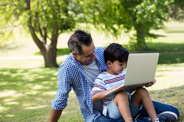 Junge, der auf dem Schoß seines Vaters sitzt und Laptop im Park benutzt