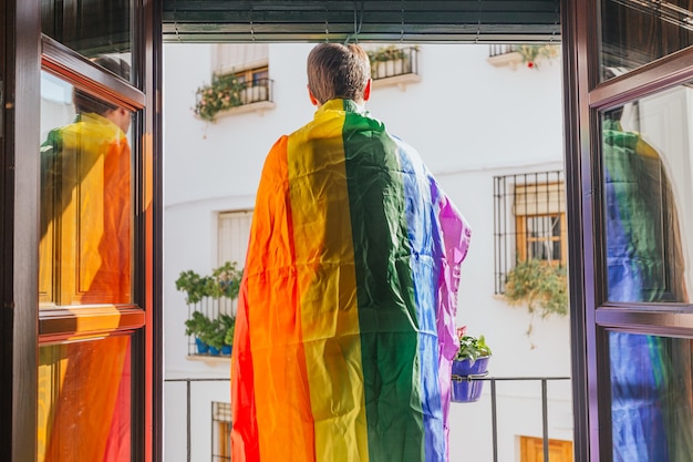 Junge, der auf einem Balkon gegenüber der Straße mit einer Regenbogenfahne auf seinen Schultern steht. lgtbi, schwul