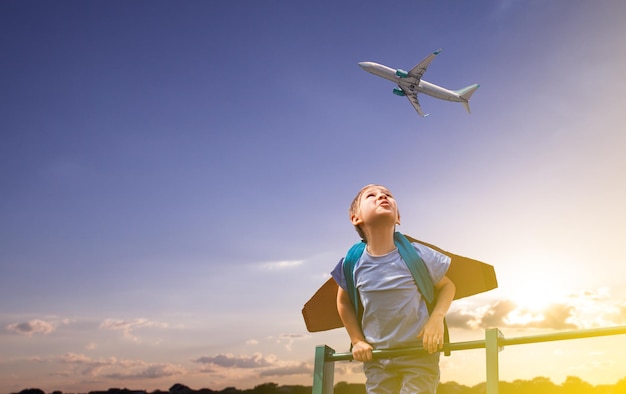 Junge, der hoch steht und das Flugzeug am Himmel betrachtet Spielplatz in der Nähe des Flughafens Lustiger kleiner Junge mit Papierflügeln auf dem Rücken, der davon träumt, Pilot zu werden