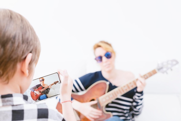 Junge, der mit dem Telefon einer Frau fotografiert, die Gitarre spielt. Selektiver Fokus