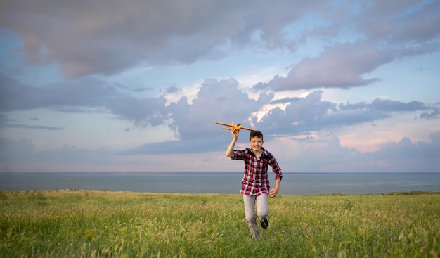 Junge, der über das Feld läuft, startet ein Spielzeugflugzeug in den Himmel bei Sonnenuntergang bewölktem Hintergrund Ein Kind geht allein und spielt in der Natur Ein Teenager-Pilot