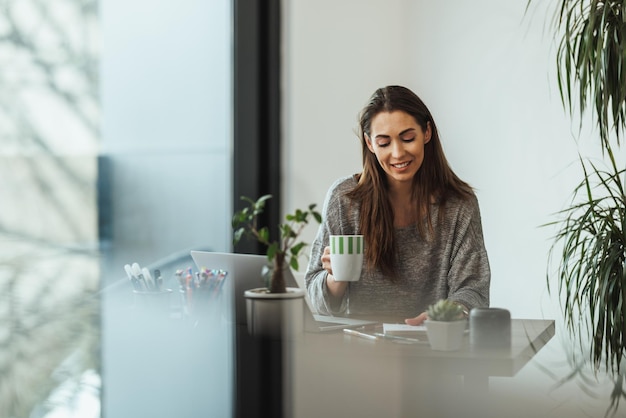 Junge Designerin trinkt Kaffee, während sie in ihrem Heimstudio arbeitet.