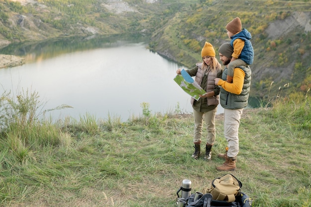 Junge dreiköpfige Familie, die sich die Karte anschaut, während sie auf Gras gegen einen Fluss oder See in natürlicher Umgebung steht und darüber nachdenkt, wohin sie als nächstes gehen sollen?