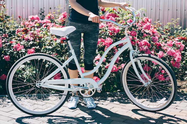 Junge dünne Frau, die mit Fahrrad auf einem Hintergrund von Büschen mit Rosen steht