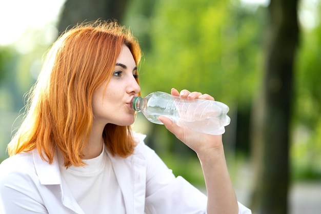 Junge durstige rothaarige Frau, die Wasser von einer Flasche im Sommerpark trinkt