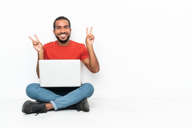 Junge ecuadorianische Mann mit einem Laptop auf dem Boden sitzend isoliert auf weißem Hintergrund mit Victory-Zeichen mit beiden Händen