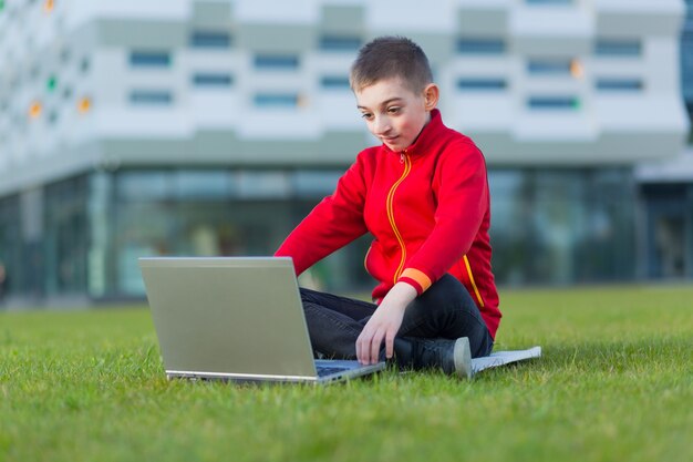 Junge, ein Schüler der Schule, der auf dem Rasen sitzt, benutzt einen Laptop