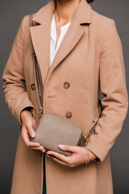 Junge elegante Dame im stilvollen braunen Mantel, der kleine Lederhandtasche hält, während auf Grau posiert